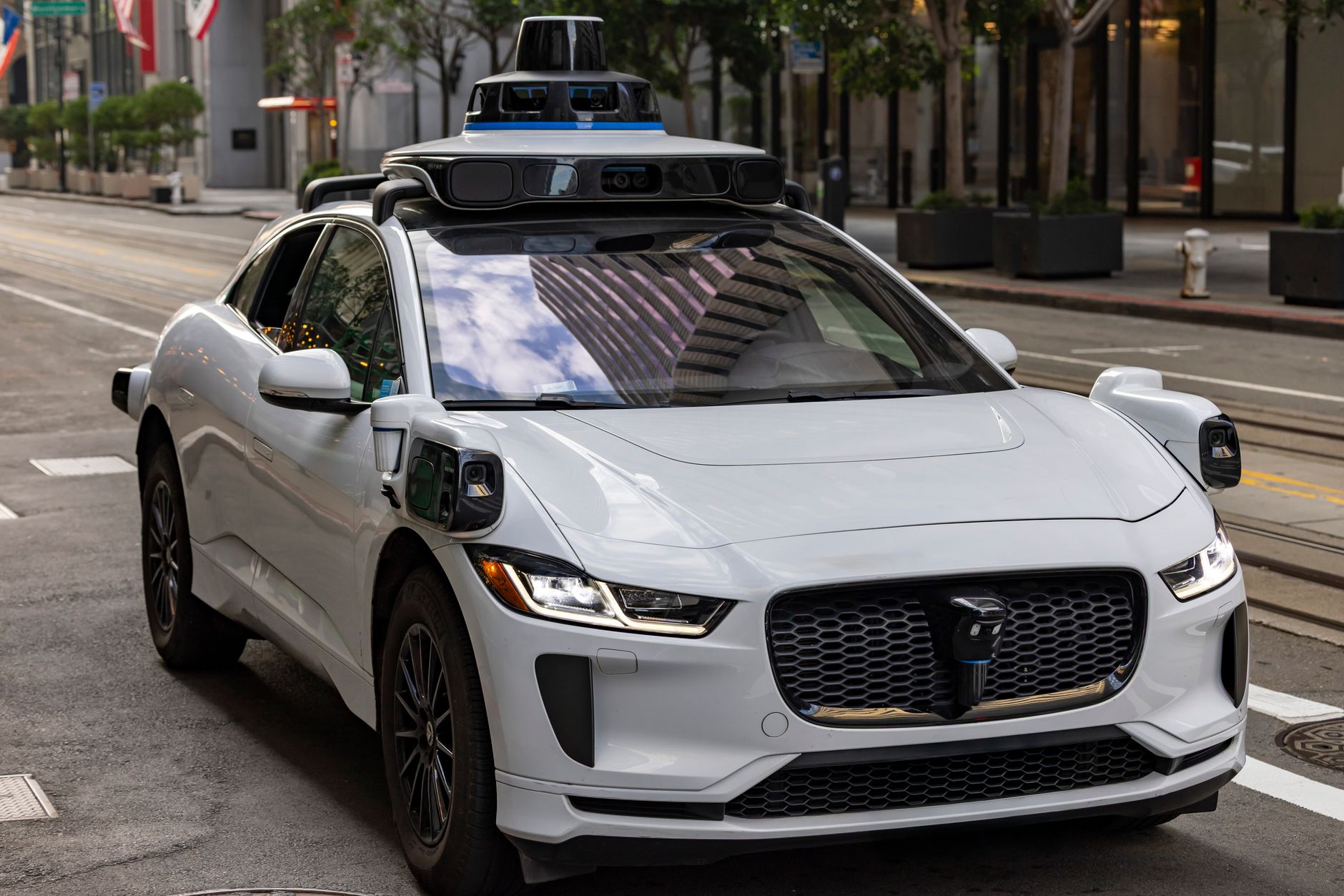 A white autonomous vehicle with roof-mounted sensors travels along a city street.