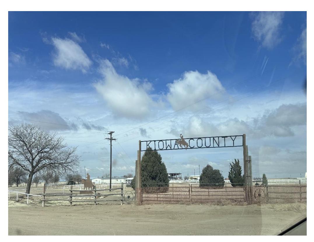 A roadside view of a fenced facility with an entrance sign reading “KIOWA COUNTY.”