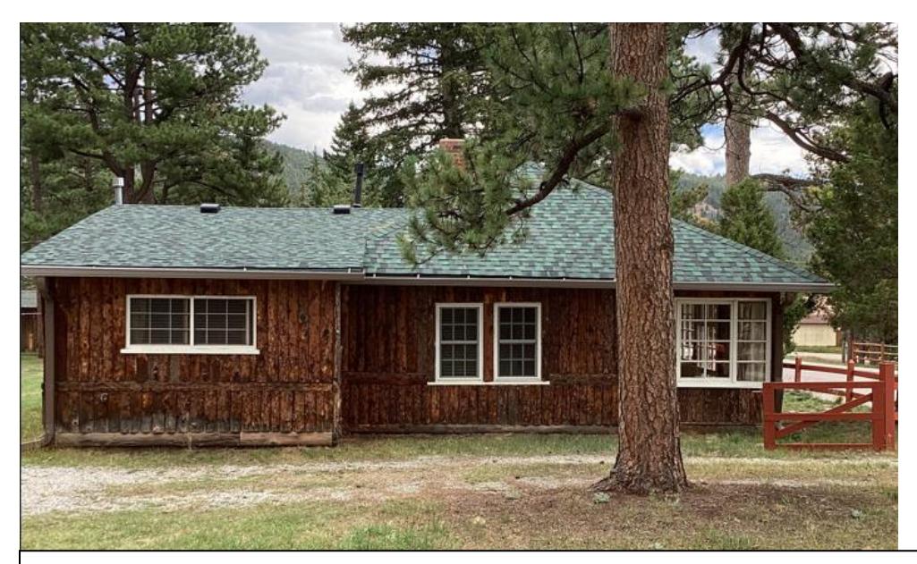 The Griffith Cabin’s west elevation, with dark wood siding, a green-shingled roof and windows; a pine trunk partly obscures the building.
