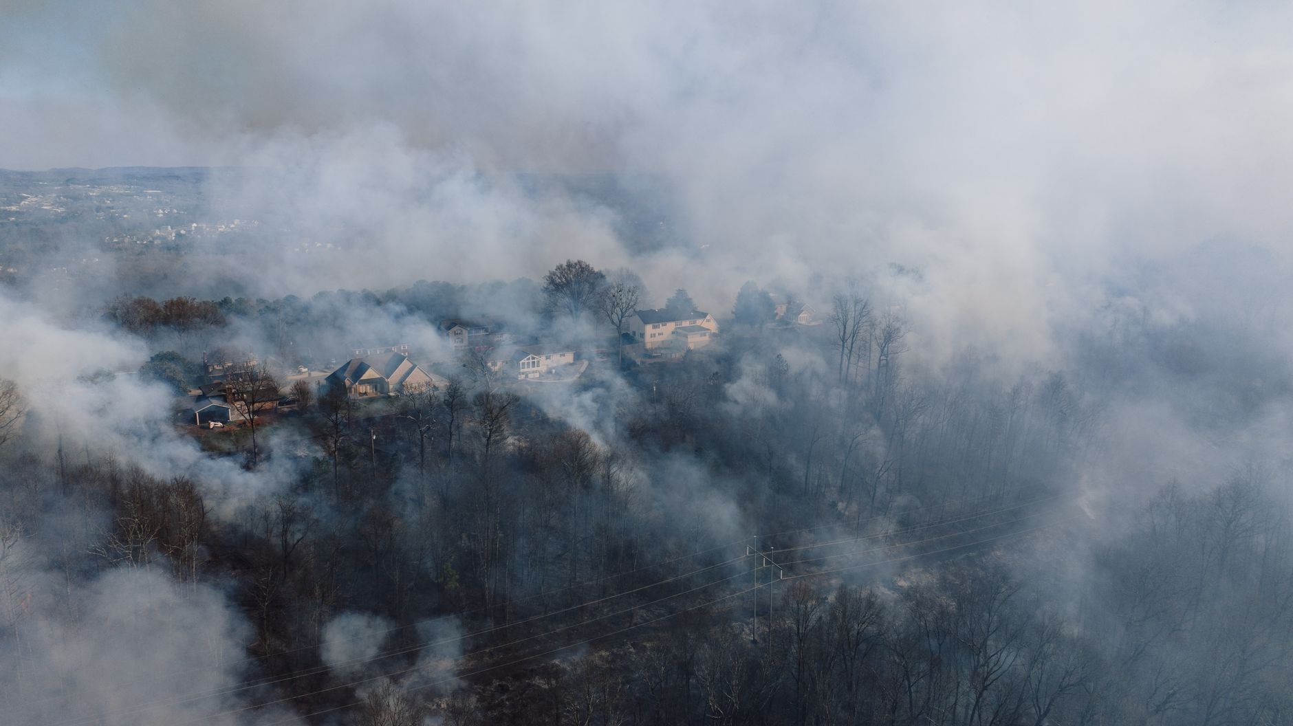 Smoke rises above homes along the edge of a wooded area.